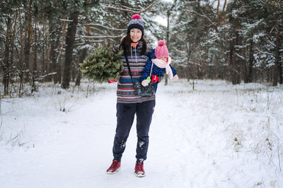 Young woman, single mom and toddler baby girl carries christmas tree in the winter park. preparing