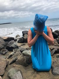 Woman wearing plastic bag while kneeling at beach against sky