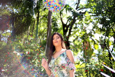Low angle portrait of beautiful woman holding colorful balloon while standing against trees in park