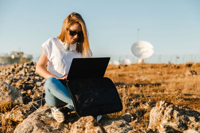 Young woman wearing sunglasses with laptop sitting on land against sky
