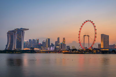 Illuminated singapore flyer and buildings by sea in city at sunset