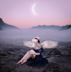 Young woman sitting on rock against mountains at sunset