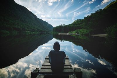 Rear view of man sitting on pier over lake by mountains against sky