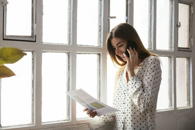 Happy young woman on cell phone looking at document at the window