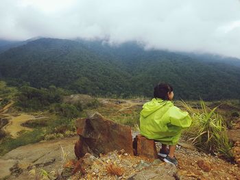 Girl sitting on rock by landscape and mountains against cloudy sky