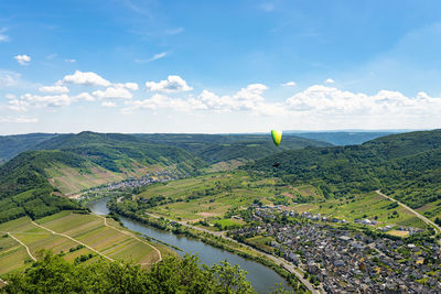 Man flying a green paraglider over beautiful wineries in germany, visible river and forest.