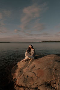 Woman sitting on rock by sea against sky during sunset