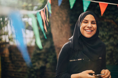 Portrait of smiling young woman in hijab holding mobile phone at backyard