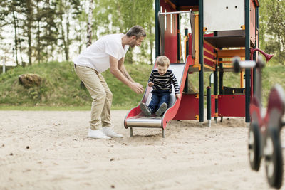 Happy father looking at son playing on slide at playground