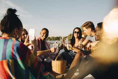 Woman photographing cheerful friends toasting drinks at jetty in summer