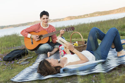Teenage girl reading book with boyfriend playing guitar during picnic