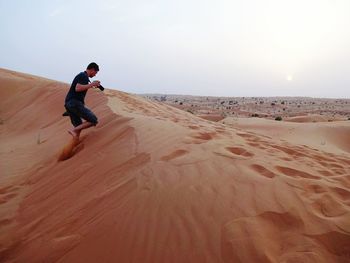 Side view of man holding camera while walking on sand dune against clear sky