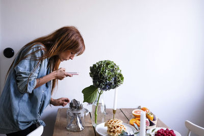 Side view of blogger photographing food at table through mobile phone by wall