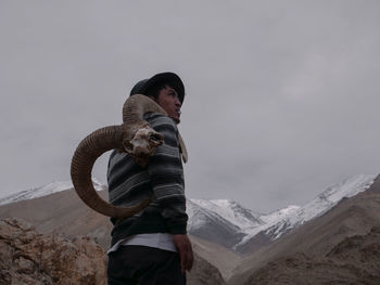 Side view of young man carrying horns while standing on mountain against cloudy sky