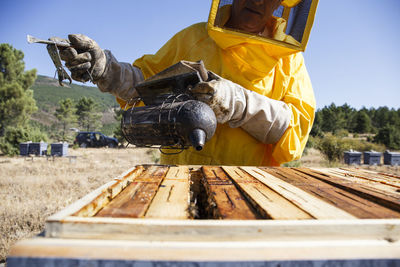 Beekeeper in protective suit using smoker on beehive