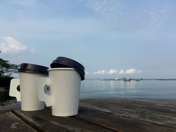 Disposable coffee cups on pier by sea against sky