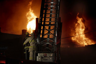 Rear view of firefighters by ladder against fire at night