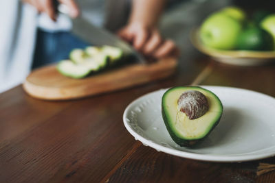 Female hands cut avocado on wooden working surface in kitchen at the home