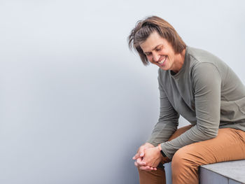 Man with long hair and happy look sits near grey wall. smiling man in earth toned casual clothes