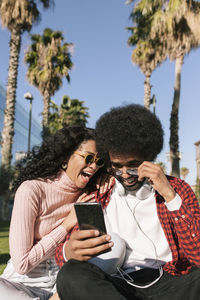 Cheerful woman sitting by man using smart phone on sunny day