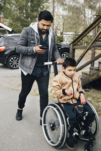 Father using smart phone while pushing autistic son sitting on wheelchair