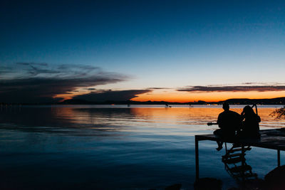 Rear view of silhouette friends sitting at pier by river against sky during sunset