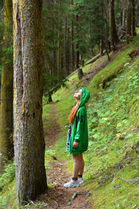 Side view of woman wearing hooded jacket standing by tree trunk in forest