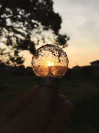 Close-up of hand holding light bulb against sky during sunset