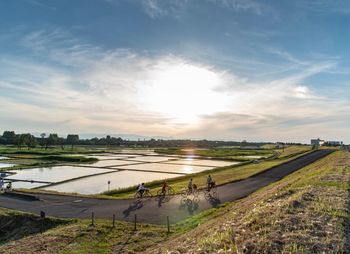 People riding bicycle by field against sky during sunny day