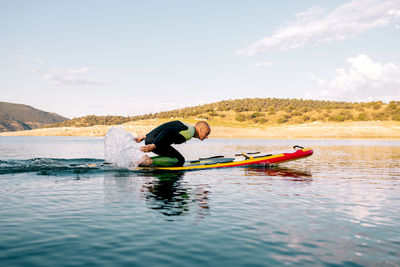 Full body side view of adult male in wetsuit kneeling on paddle board and paddling with arms on calm lake water surface