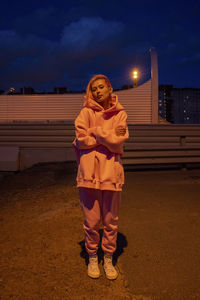Portrait of young woman with pink hair wearing track suit standing in front of building at night