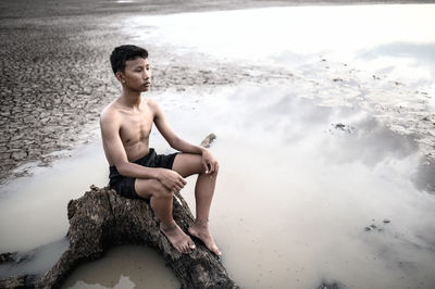 Thoughtful shirtless boy sitting on rock during arid climate