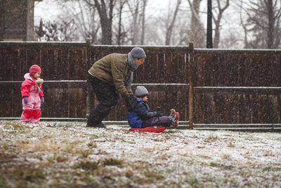 Daughter looking at father tobogganing son on snow covered field at yard during snowfall