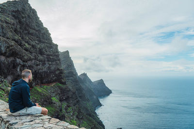 Rear view of man sitting on rock by sea against mountain