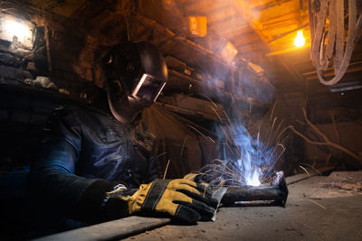 Young mechanic with a welding machine in an old dirty garage at night. man in protection doing