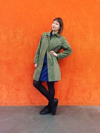 Full length portrait of woman standing with hand on hip against orange wall