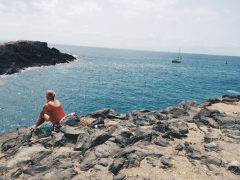 Man sitting on rock by sea against sky