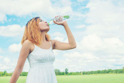 Young woman drinking water from bottle against cloudy sky