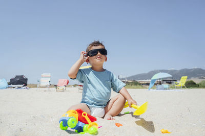 Boy adjusting sunglasses sitting with toys on sand at beach