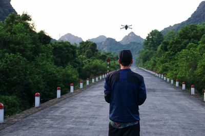 Rear view of man flying drone while standing on country road amidst trees