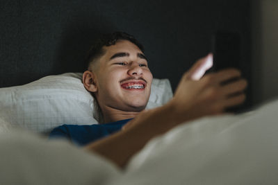 Smiling teenage boy using smart phone while lying in bedroom at home