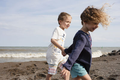 Happy siblings holding hands while walking at beach against sky during sunny day