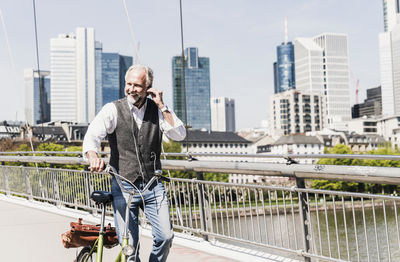 Smiling mature man with earbuds and bicycle crossing bridge in the city