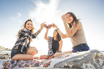 Happy friends having drink while sitting on textile at beach