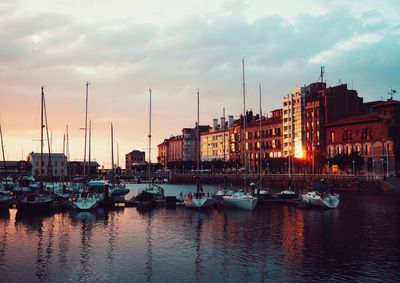 Sailboats moored on harbor in city against sky during sunset