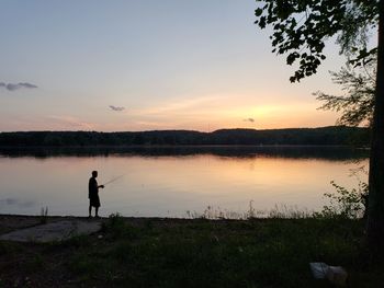 Silhouette man fishing by lake against sky during sunset