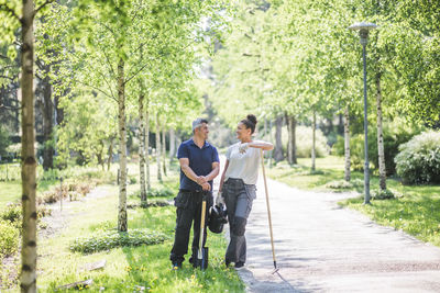 Full length of smiling female trainee looking at male instructor holding gardening equipment on footpath