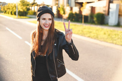 Smiling young woman gesturing peace sign while standing on road in city