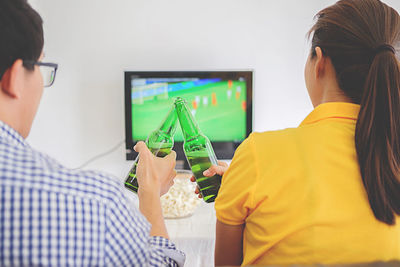Rear view of cheerful friends enjoying soccer match while holding bottle at home