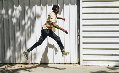 Side view of smiling african american male in stylish wear in moment of jumping near building on sunny day during city stroll
